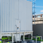 A rooftop industrial cooling tower with white metal panels and large green pipes extending from its base, highlighting an efficient cooling tower water treatment system. A blue sky and sections of a beige building with windows are visible in the background.