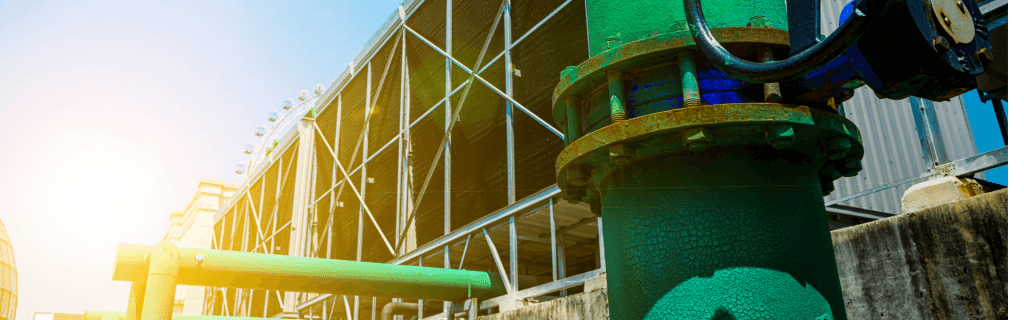 Green industrial pipes with valves in the foreground, against a backdrop of a large metal framework and cooling tower structure. The sun is setting on the left, casting a warm glow across the scene, while water treatment processes silently ensure efficiency and sustainability.