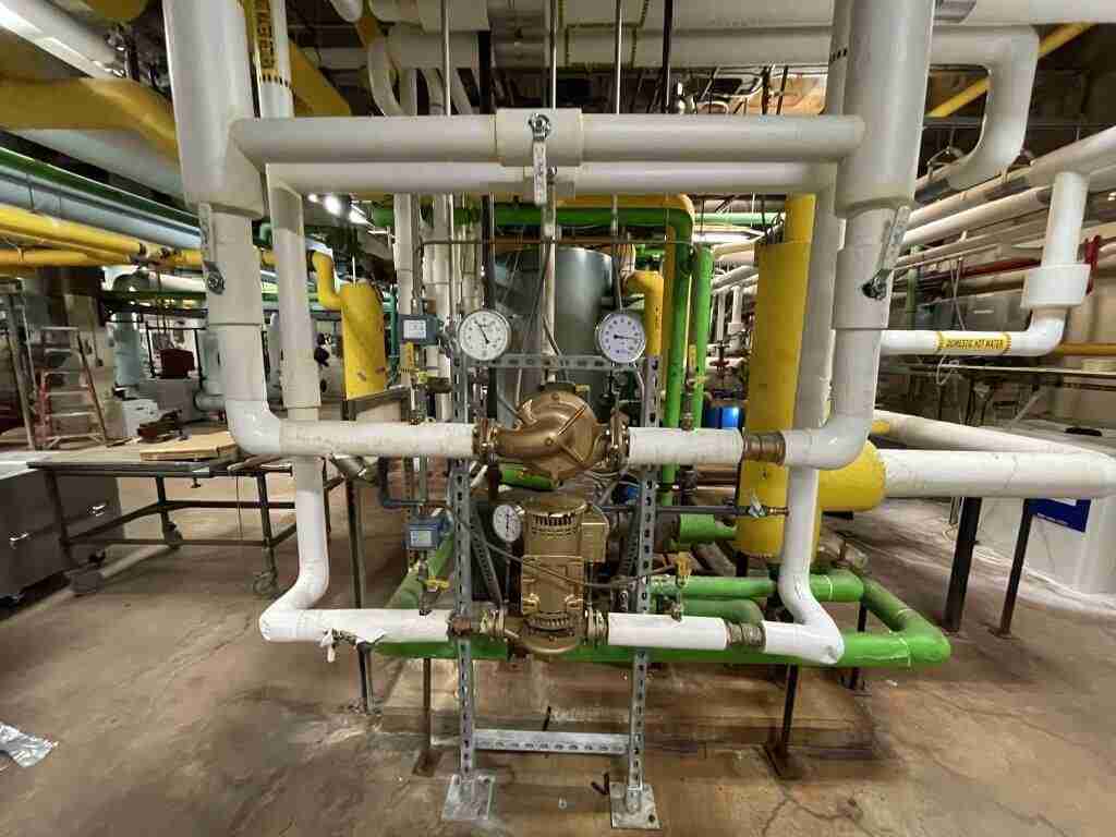 Influent Water Treatment, A complex network of white pipes and gauges on an industrial gas meter assembly hints at the precision found in commercial water treatment systems. Two pressure gauges display readings, while pipes are connected by brass fittings, with green and yellow elements visible in the background.