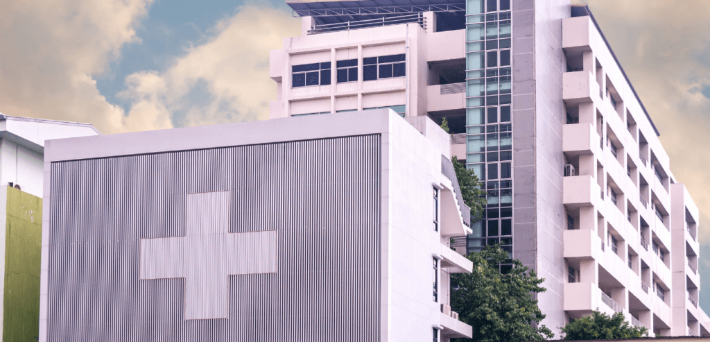 A modern multi-story building with a large white cross on the facade, likely a healthcare facility, stands under a partly cloudy sky. Nearby, trees sway gently in the breeze, adding a touch of nature to the scene.
