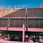 Massive cooling towers with metal gratings stand outdoors under a clear blue sky. Pipes and valves are visible at the base, supported by sturdy structural beams.