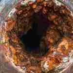 Close-up of a severely corroded and rusted metal pipe with a large hole in the center. The surrounding metal shows flaking and discoloration from galvanic corrosion, indicating significant deterioration over time.