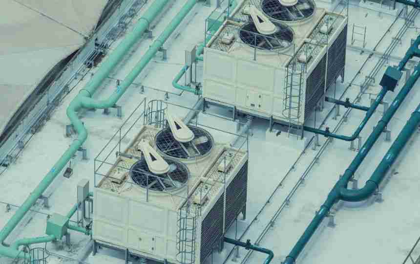 Two large industrial cooling tower units with visible fans and vents are mounted on a rooftop. A network of green and blue pipes surrounds them, designed to optimize approach temperature. The top-down perspective offers a clear view of the equipment layout.