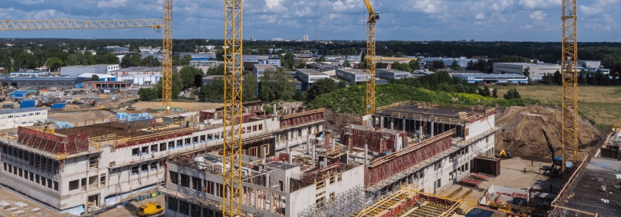 A bustling construction site features several large cranes erecting a multi-story structure. Surrounded by industrial buildings and greenery, scaffolding and construction materials are visible. Water management systems are integrated seamlessly under the partly cloudy sky overhead.