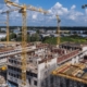 A bustling construction site features several large cranes erecting a multi-story structure. Surrounded by industrial buildings and greenery, scaffolding and construction materials are visible. Water management systems are integrated seamlessly under the partly cloudy sky overhead.