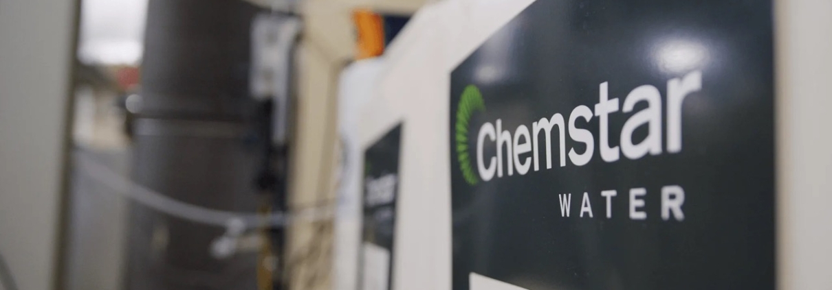 Close-up of a sign on a wall with the words Chemstar Water in white letters on a dark background. The image, slightly blurred on the left side, reveals some Cooling Tower Water Treatment equipment or machinery in the background.