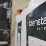 Close-up of a sign on a wall with the words Chemstar Water in white letters on a dark background. The image, slightly blurred on the left side, reveals some Cooling Tower Water Treatment equipment or machinery in the background.