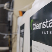 Close-up of a sign on a wall with the words Chemstar Water in white letters on a dark background. The image, slightly blurred on the left side, reveals some Cooling Tower Water Treatment equipment or machinery in the background.