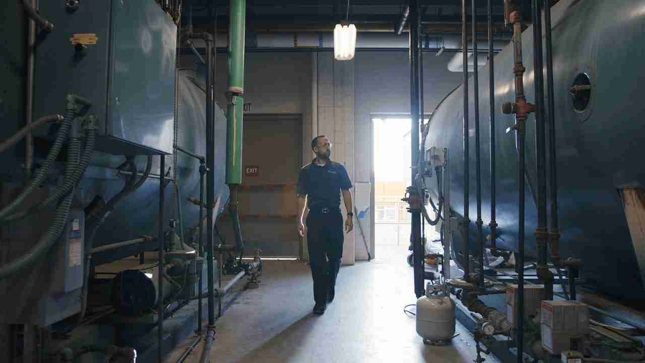 A confident Chemstar WATER technician walks between two large chillers in a mechanical room of a large institution, ensuring optimal system performance.