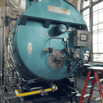 A large industrial teal-blue boiler labeled Cleaver Brooks is indoors. It features various pipes, gauges, and a control panel. A red ladder stands nearby. Large windows in the background allow natural light in.