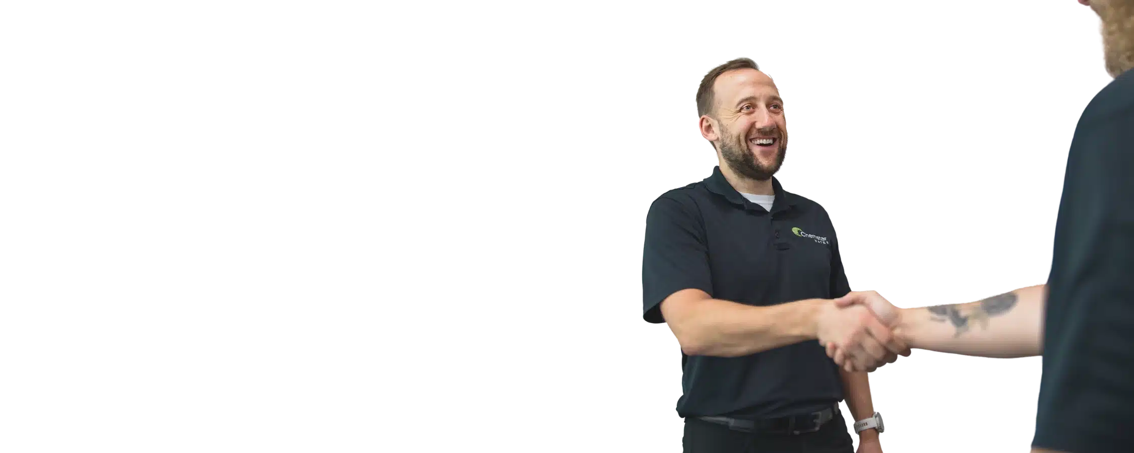A smiling man in a black polo shirt shakes hands with another person, whose arm and partial beard are visible in the foreground, against a plain background.