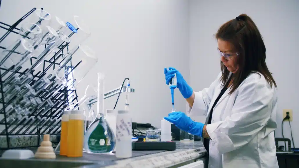 A scientist in a lab coat and safety glasses uses a pipette at a laboratory bench, surrounded by flasks, beakers, and laboratory glassware filled with various liquids.