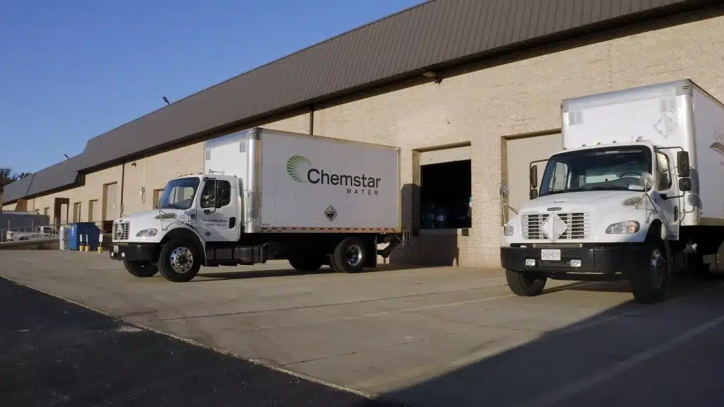 Two white delivery trucks, both labeled Chemstar WATER, are parked outside a beige industrial building with garage doors on a sunny day.