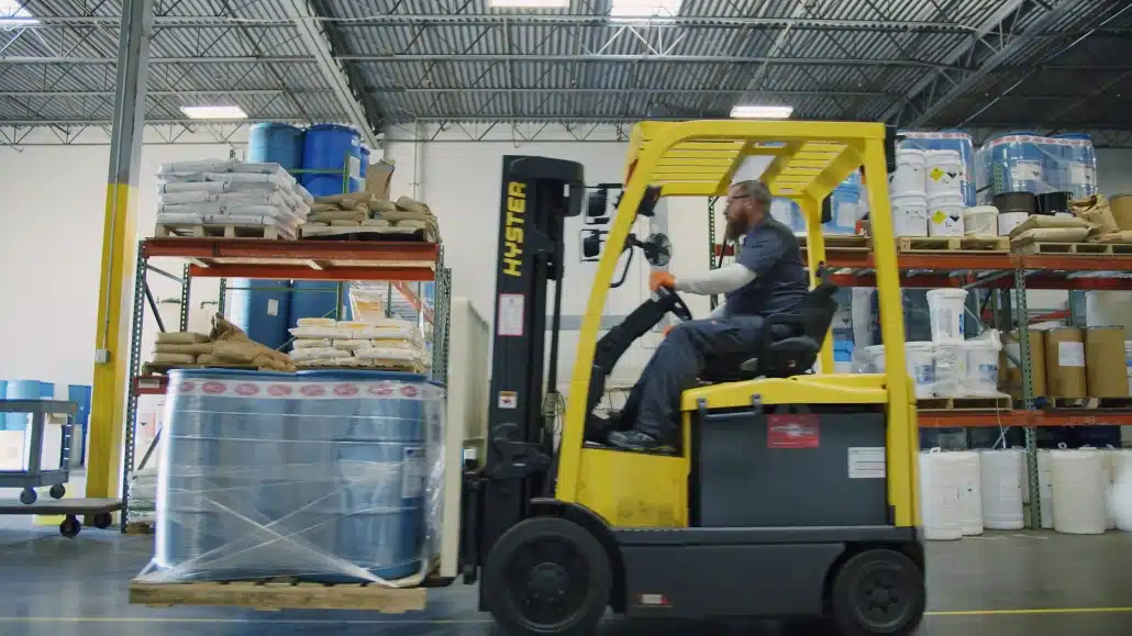 A worker operates a yellow forklift inside a warehouse, moving a pallet wrapped in plastic. Shelves stacked with bags and blue barrels are visible in the background under a metal roof.