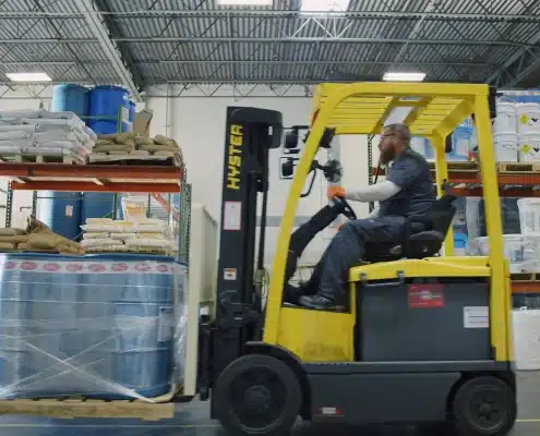 A worker operates a yellow forklift inside a warehouse, moving a pallet wrapped in plastic. Shelves stacked with bags and blue barrels are visible in the background under a metal roof.