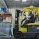 A worker operates a yellow forklift inside a warehouse, moving a pallet wrapped in plastic. Shelves stacked with bags and blue barrels are visible in the background under a metal roof.