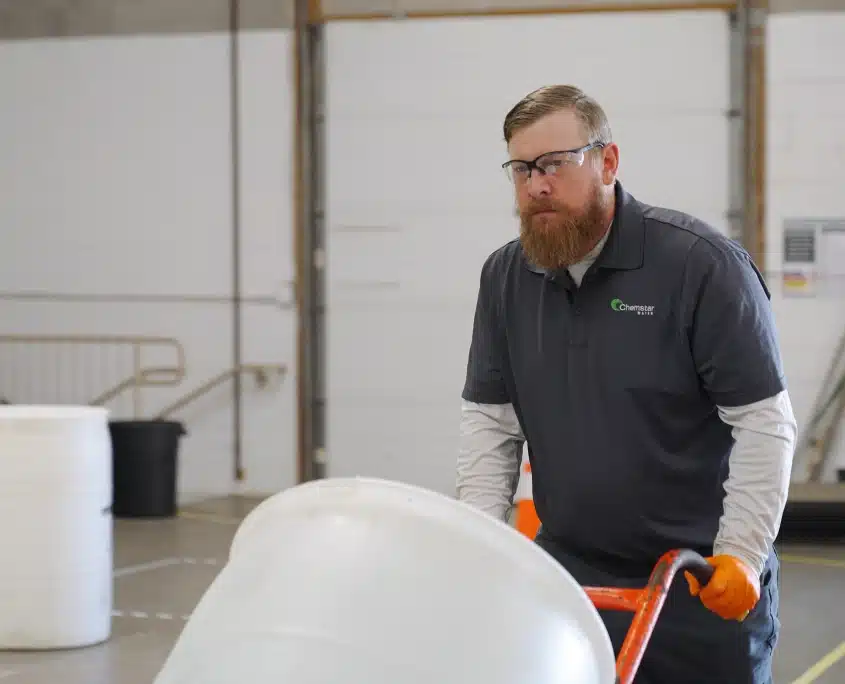 Government and Military Water Treatment Services | A man with a beard wearing safety glasses, gloves, and a dark polo shirt uses a hand truck to move a large white barrel inside an industrial warehouse. Other barrels are visible in the background.