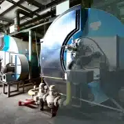 Two large industrial boilers in a modern facility, surrounded by pipes, valves, and pumps, with metal walls and overhead lighting visible in the background.