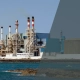 A large industrial desalination plant with multiple smokestacks stands by the sea. The left side is clear and colorful, while the right side is dimmed with a grey overlay. Several boats are visible on the water.