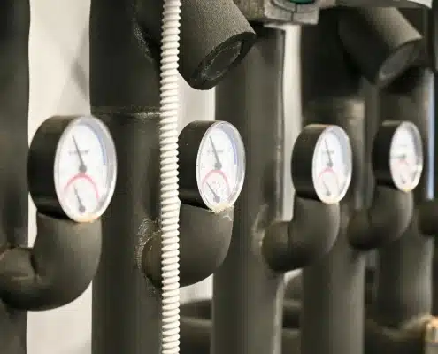 Close-up of several industrial pressure gauges attached to insulated black pipes, with coiled white cables running alongside them. The gauges display measurements and are aligned in a row.