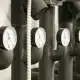 Close-up of several industrial pressure gauges attached to insulated black pipes, with coiled white cables running alongside them. The gauges display measurements and are aligned in a row.