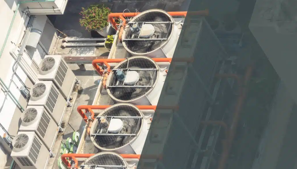 Aerial view of large industrial air conditioning units with visible fans and orange pipes on a rooftop, surrounded by smaller AC units and potted plants.
