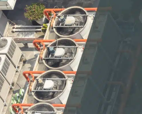 Aerial view of large industrial air conditioning units with visible fans and orange pipes on a rooftop, surrounded by smaller AC units and potted plants.