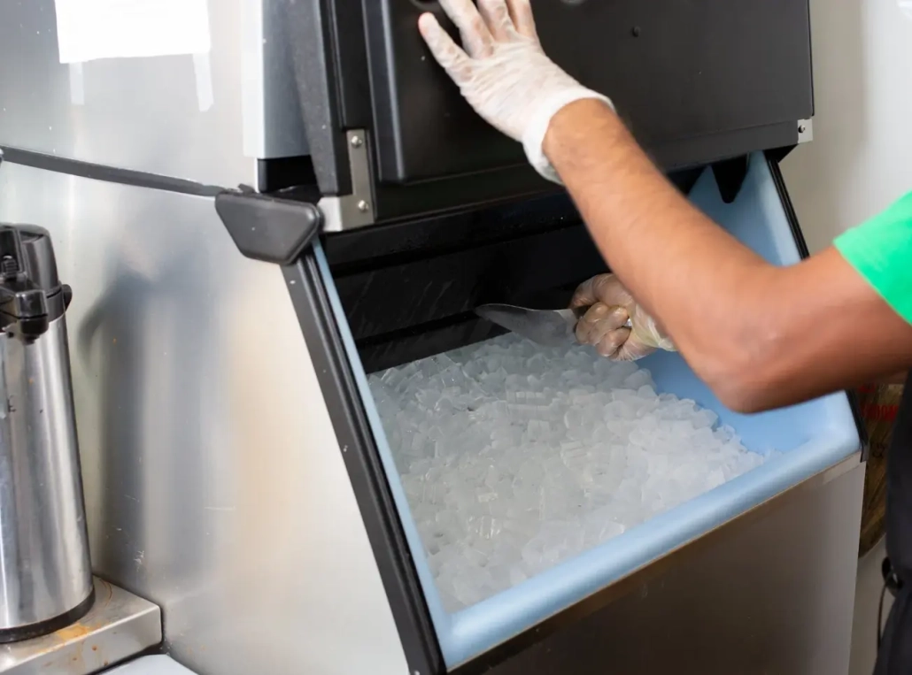 A person wearing gloves scoops ice from a commercial ice machine, lifting the lid with one hand and holding a scoop in the other—highlighting the importance of regular Commercial Ice Machine Cleaning & Legionella Prevention for safety.