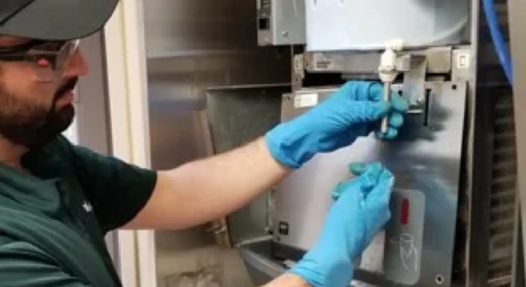 Wearing blue gloves and safety glasses, a person uses both hands to service equipment, inserting or removing a component from a metallic machine with a visible valve and tubing for commercial ice machine cleaning & Legionella prevention.
