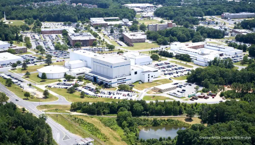 Aerial view of the NASA Goddard Space Flight Center campus, featuring multiple white buildings, parking lots, and surrounding green trees, with a road and wooded area in the foreground.