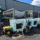 Several large industrial cooling units are lined up outdoors with white plastic water tanks, hoses, pumps, and a portable generator in front of a gray building under a blue sky with clouds.