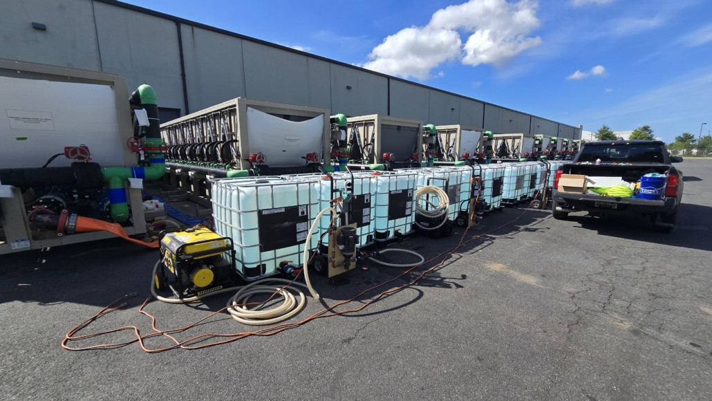 Large white chemical tanks connected by hoses and machinery are lined up outside an industrial building, ready for cooling tower cleaning. A black pickup truck with equipment in its bed is parked nearby under a blue sky with scattered clouds.