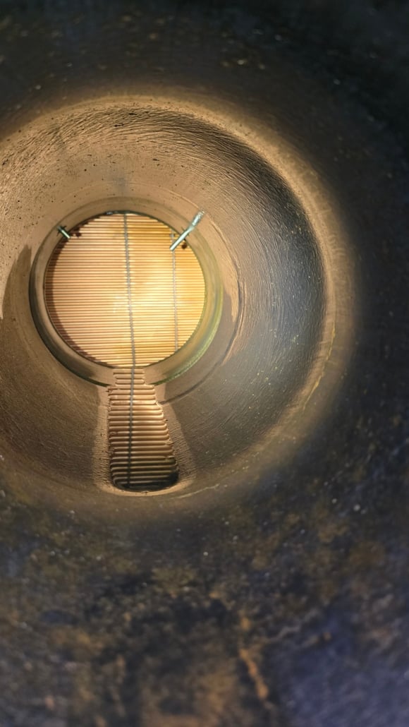 View from inside a large, cylindrical metal pipe, often seen in cooling tower cleaning, looking toward the grated metal gate at the far end, illuminated by warm light. The pipe’s inner surface appears textured and slightly worn.