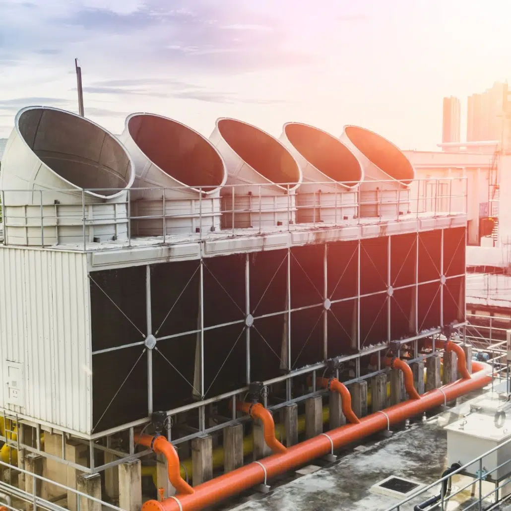 A row of large industrial cooling towers with circular vents and orange pipes is situated on a rooftop in an urban area, illuminated by afternoon sunlight, highlighting the importance of Legionella prevention and proper cooling tower water treatment. A row of large industrial cooling towers with circular vents and orange pipes is situated on a rooftop in an urban area, illuminated by afternoon sunlight, highlighting the importance of Legionella prevention and proper cooling tower water treatment.