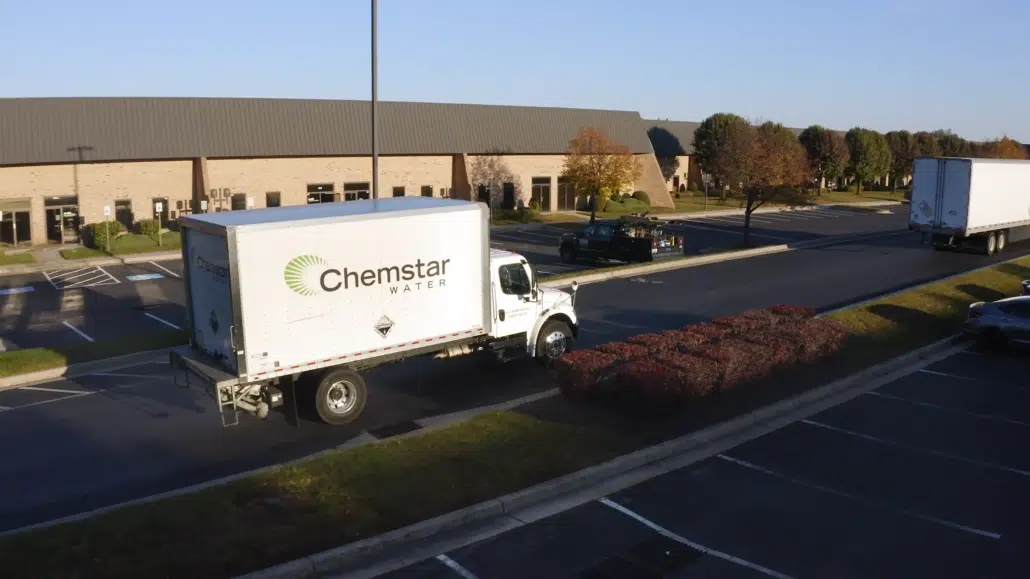 A white Chemstar Water delivery truck promoting Microbiological Control is parked near a low hedge in a nearly empty office parking lot on a clear day, with a brick building and a few trees in the background. A white Chemstar Water delivery truck promoting Microbiological Control is parked near a low hedge in a nearly empty office parking lot on a clear day, with a brick building and a few trees in the background.