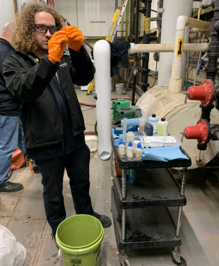 A person with curly hair and orange gloves examines a sample in a lab setting near pipes. There is a cart with bottles, a green bucket, and industrial equipment in the background.