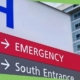 A hospital sign with a large blue H and arrows for Emergency and South Entrance stands before a modern healthcare facility, where effective water management and Legionella prevention are crucial to patient safety.